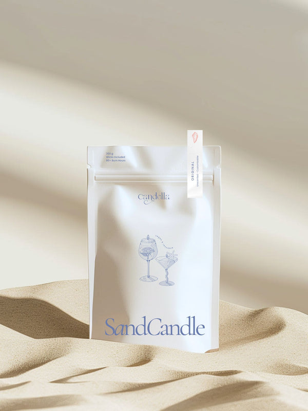 A product image of Candella's Sand Candle. The minimalist, cream-coloured pouch features a subtle illustration of two cocktail glasses with text that reads "Vegan & Reusable." A small label at the top right corner of the package indicates the scent "Original- Unscented & Customizable"