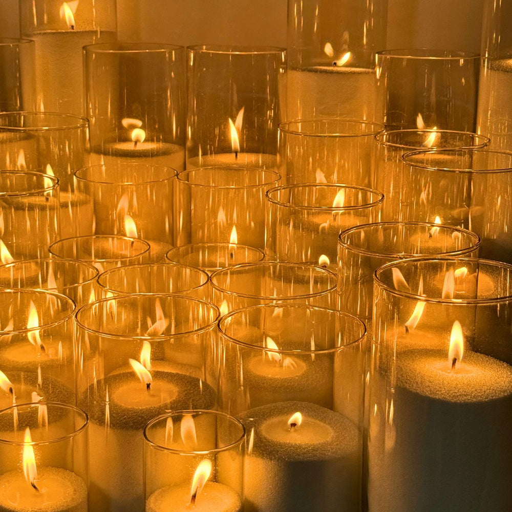 
                      
                        A display of multiple sand candles burning inside clear glass cylinders of various heights, arranged in rows. Each candle has a soft, warm flame, creating an ambient glow against a neutral background. 
                      
                    