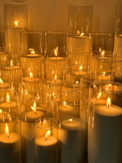 A display of multiple sand candles burning inside clear glass cylinders of various heights, arranged in rows. Each candle has a soft, warm flame, creating an ambient glow against a neutral background. 