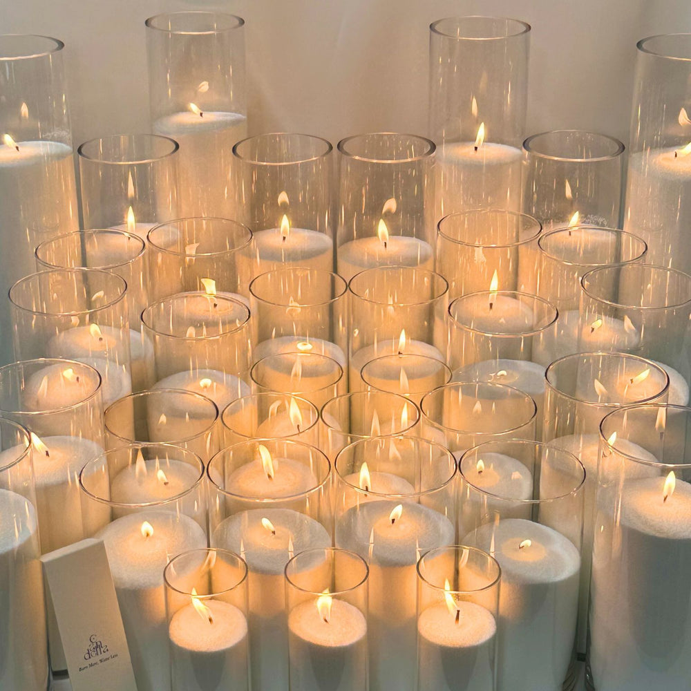 A collection of sand candles made from a 25kg bag of wax, set inside tall, clear glass cylinders of varying sizes, with each candle emitting a warm flame, showcasing the product's capacity for creating multiple candles.