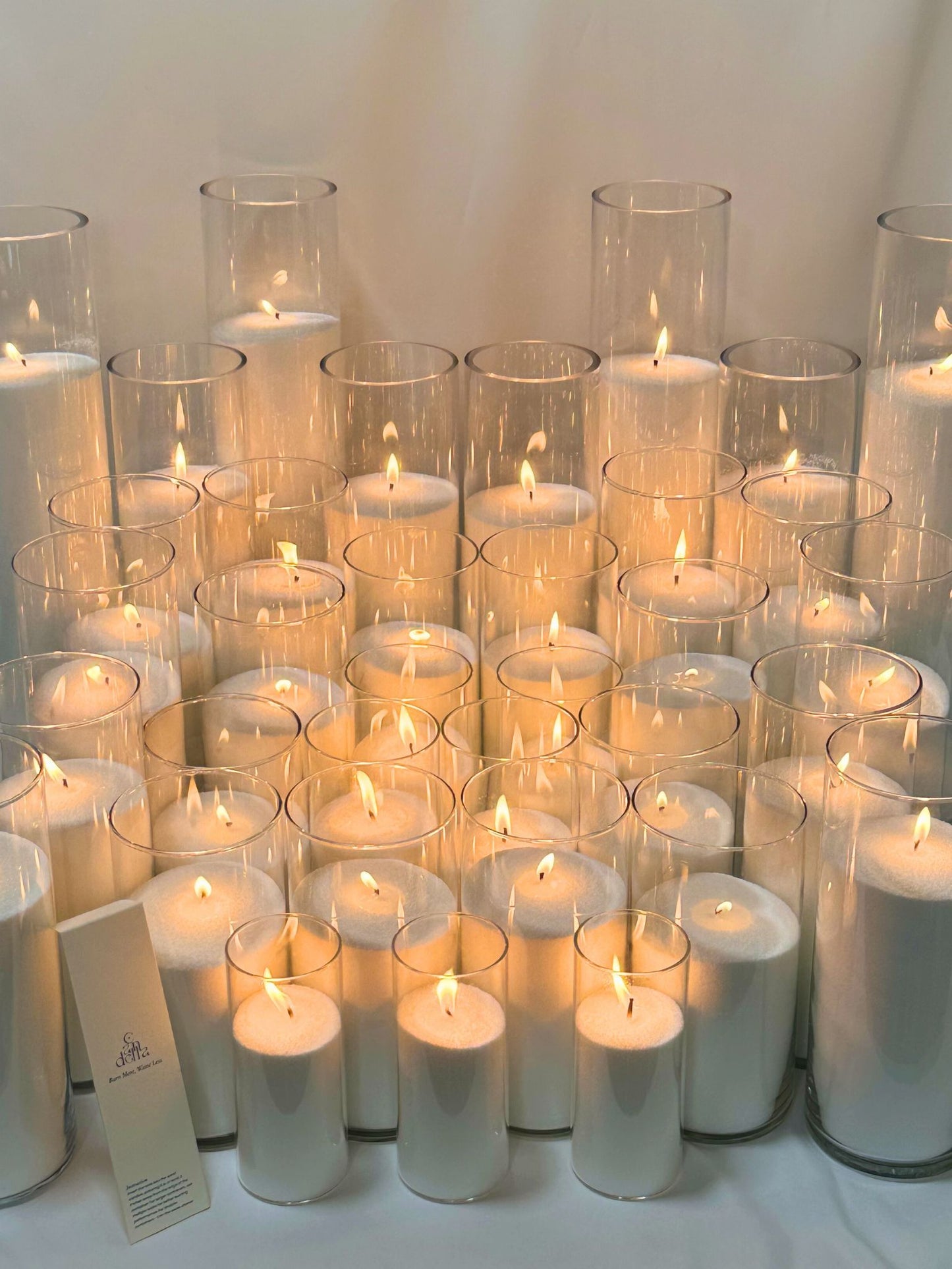 A collection of sand candles made from a 25kg bag of wax, set inside tall, clear glass cylinders of varying sizes, with each candle emitting a warm flame, showcasing the product's capacity for creating multiple candles.