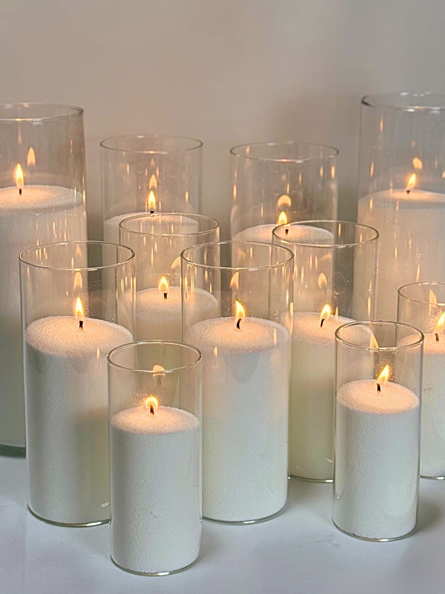 A collection of sand candles made from a 5kg bag of wax, set inside tall, clear glass cylinders of varying sizes, with each candle emitting a warm flame, showcasing the product's capacity for creating multiple candles.