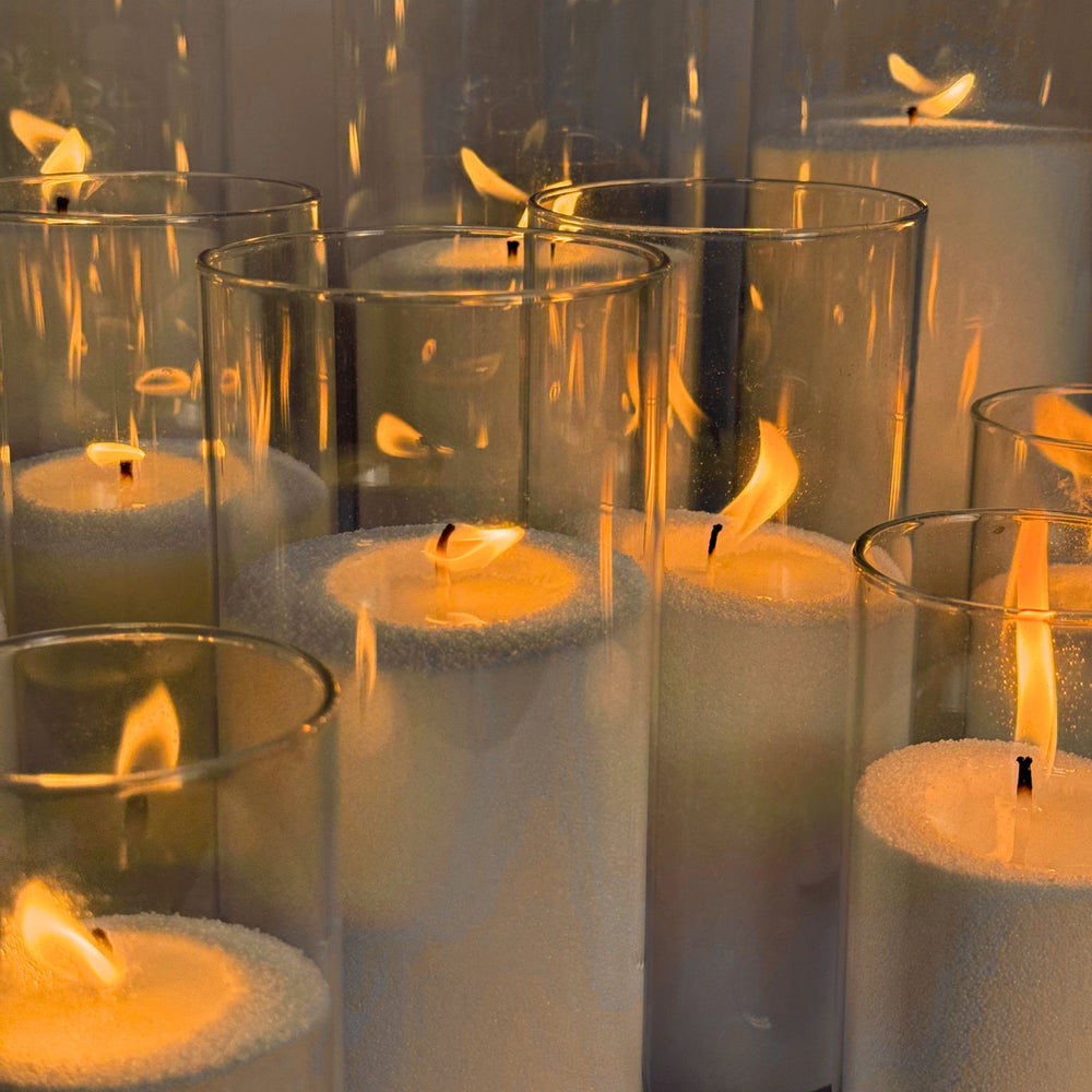 
                      
                        A display of multiple sand candles burning inside clear glass cylinders of various heights, arranged in rows. Each candle has a soft, warm flame, creating an ambient glow against a neutral background. 
                      
                    