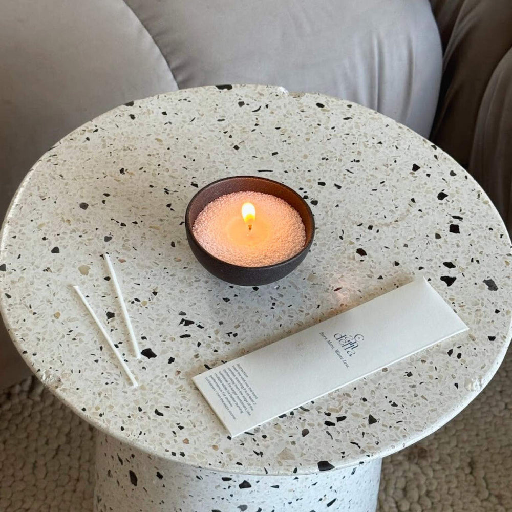 A sand candle lit on a coffee table with a candella wick bag beside it.