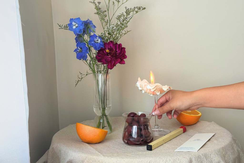 A seashell-shaped maritnin candle made with Candellas sand, surrounded by flowers, olives, oranges, lighters, and a Candellas wick bag.