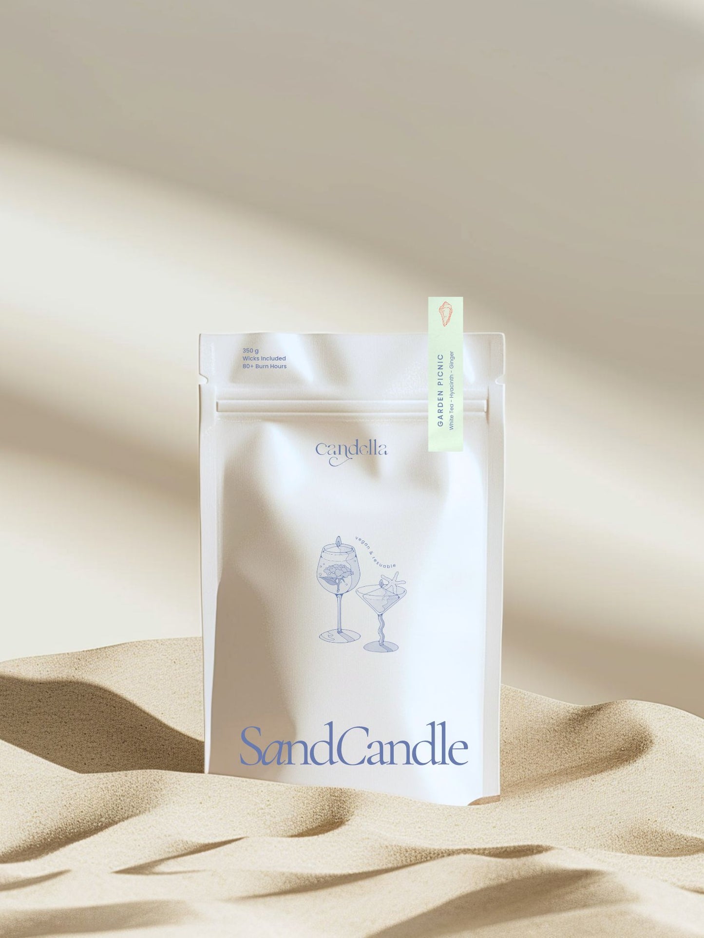 A product image of Candella's Sand Candle. The minimalist, cream-coloured pouch features a subtle illustration of two cocktail glasses with text that reads "Vegan & Reusable." A small label at the top right corner of the package indicates the scent "GARDEN PICNIC" with notes of "White Tea - Hyacinth - Ginger."