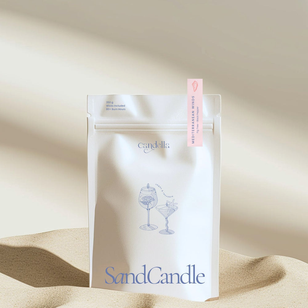 A product image of Candella's Sand Candle. The minimalist, cream-coloured pouch features a subtle illustration of two cocktail glasses with text that reads "Vegan & Reusable." A small label at the top right corner of the package indicates the scent "Mediterranean Winds" with notes of "Fig Tree - Black Pepper".