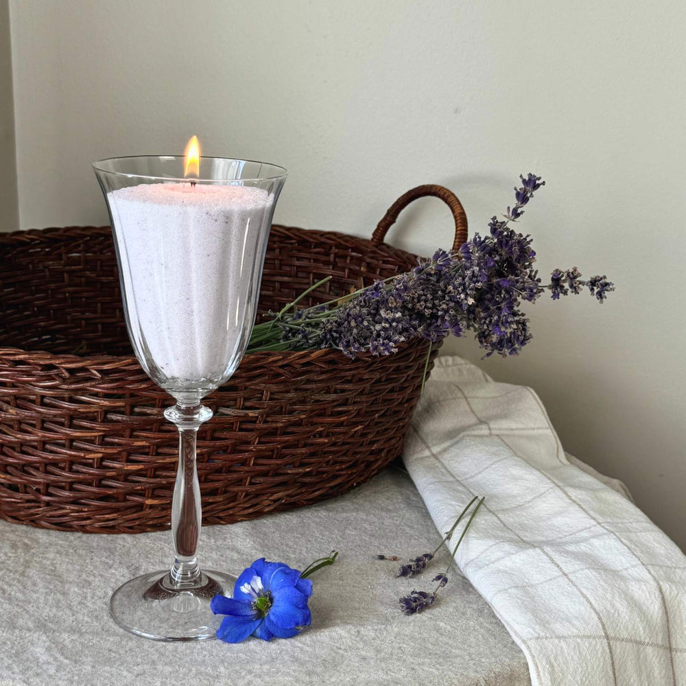 A pastel purple sand candle lit beside lavender sprigs, a basket, and flowers, made with a Lavender Fog scent from candella sand candle.