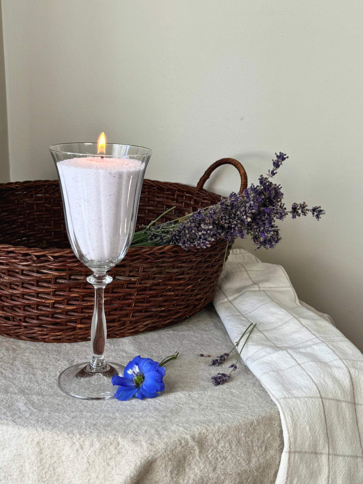 A pastel purple sand candle lit beside lavender sprigs, a basket, and flowers, made with a Lavender Fog scent from candella sand candle.